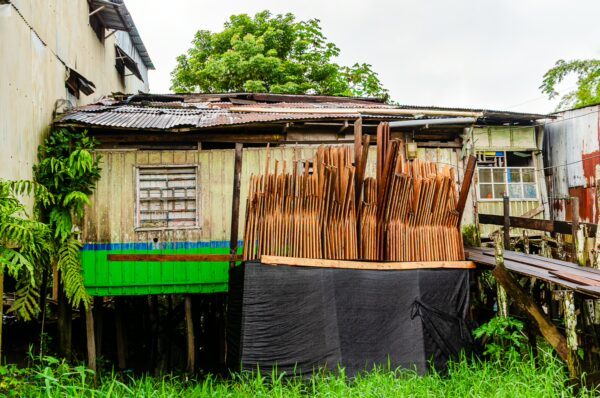 Rustic wooden house in Leticia, Amazonas, Colombia, with stacked lumber and greenery surrounding the structure.