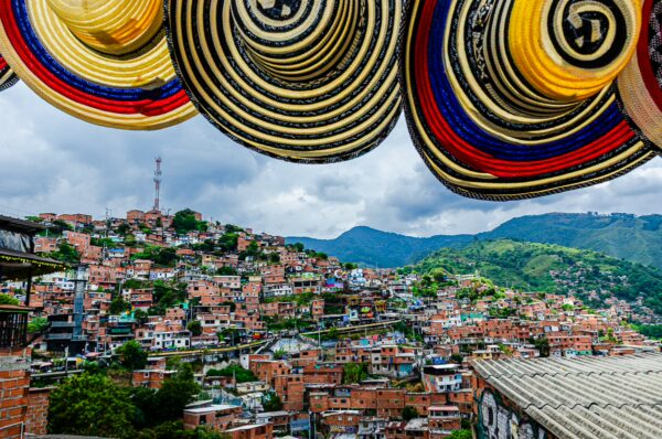Colorful hats hang overhead, framing a vibrant view of Comuna 13's hillside homes and green mountains in Medellín, Colombia.