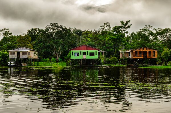 Colorful stilt houses reflect on the still waters of the Amazonas near Leticia, surrounded by lush green foliage and cloudy skies.