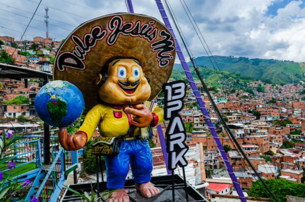 A colorful sculpture of a smiling character with a hat holding a globe, set against the vibrant hills of Comuna 13, Medellín.