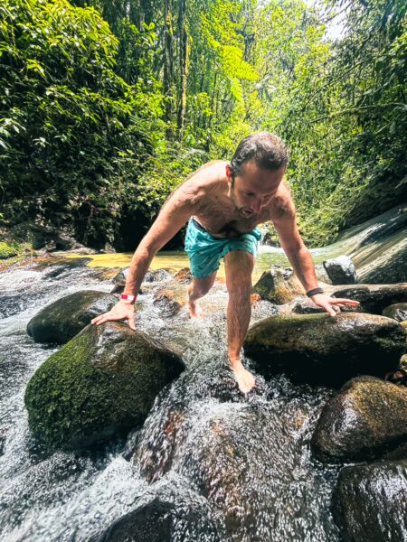 A man climbs over mossy rocks in the rushing water of Santa Rita Waterfalls, surrounded by lush greenery in Salento, Quindio, Colombia.