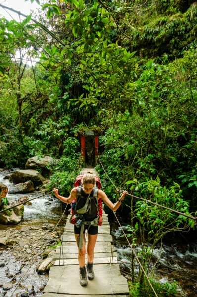 Hiker crossing a narrow bridge over a stream surrounded by lush greenery in Cocora Valley, Quindio, Colombia.