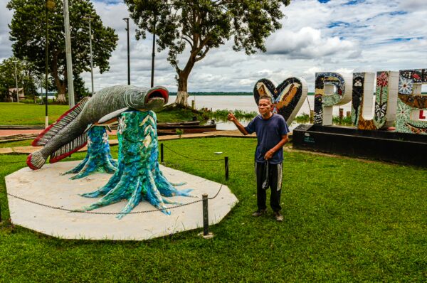 Colorful sculpture of a fish and trees in Puerto Nariño, Amazonas, Colombia, with a person standing beside it on grassy land.