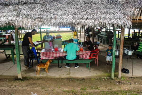 Colorful tables under a thatched roof in Puerto Narino, Amazonas, Colombia, with people enjoying a meal and a dog nearby.