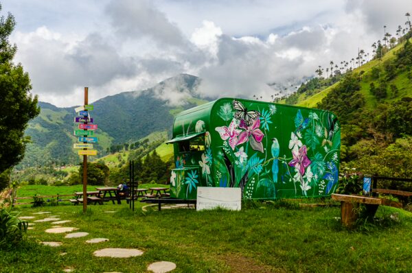 Colorful green food truck adorned with flowers, nestled in lush Cocora Valley, Quindio, Colombia, surrounded by rolling hills.