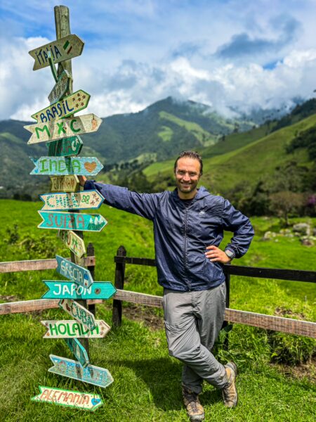 Colorful directional signs mark various destinations, with lush green hills of Cocora Valley in Quindío, Colombia, in the background.