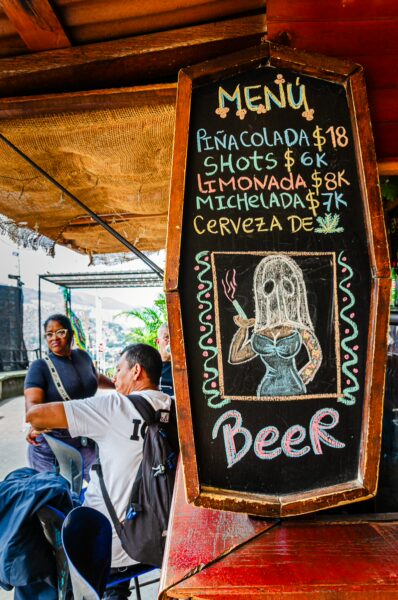 Colorful menu board featuring drinks like piña colada and michelada, with patrons in a vibrant Medellín eatery.