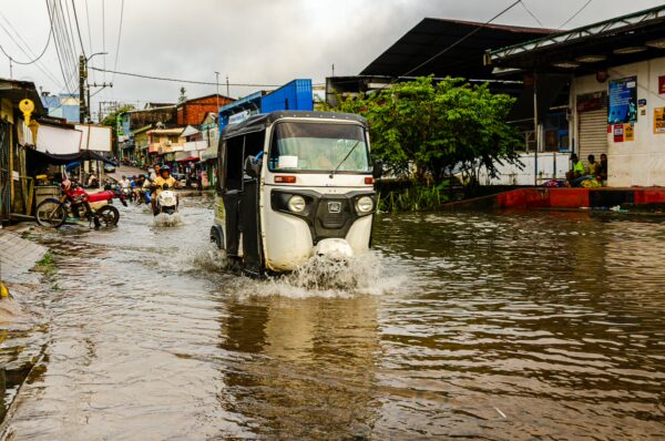 Flooded street in Leticia, Amazonas, Colombia, with a vehicle navigating water, surrounded by rain-soaked buildings and greenery.