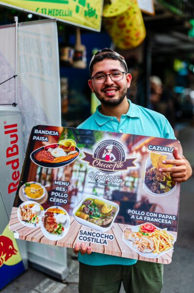 A cheerful vendor in Comuna 13, Medellín, displays a colorful sign showcasing local dishes like bandeja paisa and sancocho.