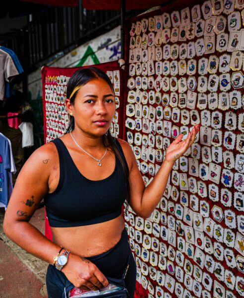 A young woman stands in Comuna 13, Medellín, showcasing colorful handmade crafts at a vibrant street market.