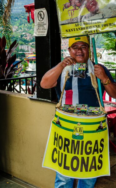 A vendor in Comuna 13, Medellín, holds a colorful sign and bucket promoting local snacks, with lush greenery in the background.
