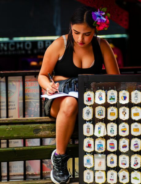 A woman with a flower in her hair sits writing beside a display of colorful souvenirs in Comuna 13, Medellín.