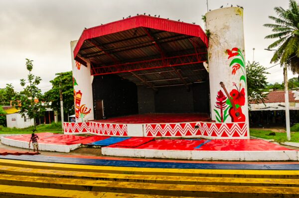 Colorful stage with decorative walls and a red roof in Leticia, Amazonas, Colombia, surrounded by greenery and striped pavement.