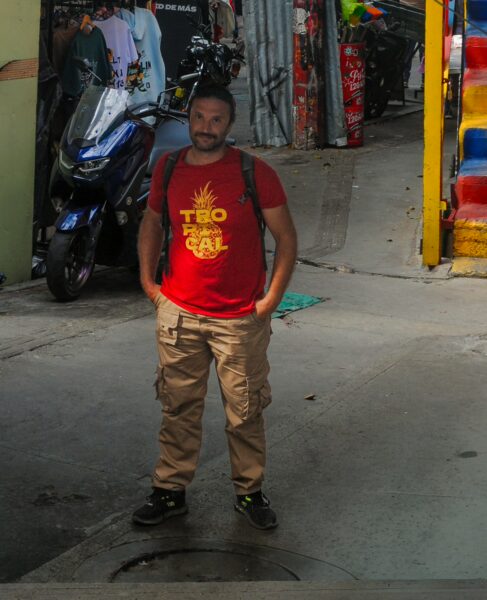 A man in a red shirt stands in a vibrant alley of Comuna 13, Medellín, surrounded by colorful street art and scooters.
