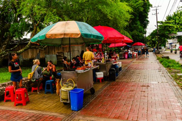 Colorful food stalls with umbrellas line the sidewalk at Parque Santander in Leticia, Amazonas, Colombia, bustling with visitors.