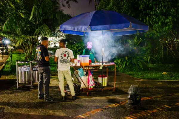 Street vendors under a blue umbrella serve food at Parque Santander in Leticia, Amazonas, Colombia, surrounded by lush greenery.