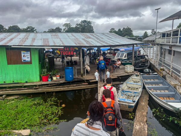 Travelers walk towards a vibrant green building along the dock in Leticia, Amazonas, Colombia, surrounded by boats and lush vegetation.