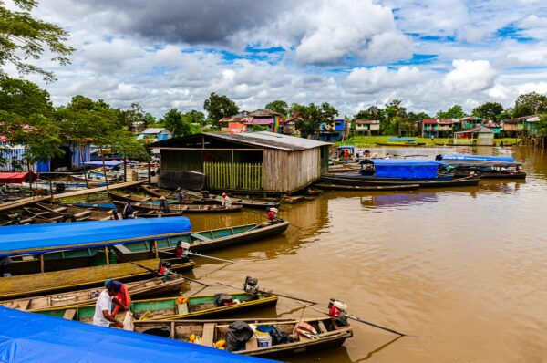Colorful boats with blue tarps line the muddy riverbank in Leticia, Amazonas, Colombia, against a backdrop of lush greenery and sky.