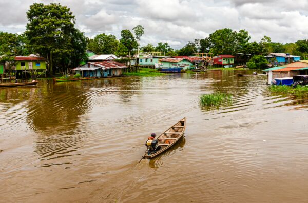 A man paddles a wooden canoe on the brown waters near colorful houses in Leticia, Amazonas, Colombia, surrounded by lush greenery.