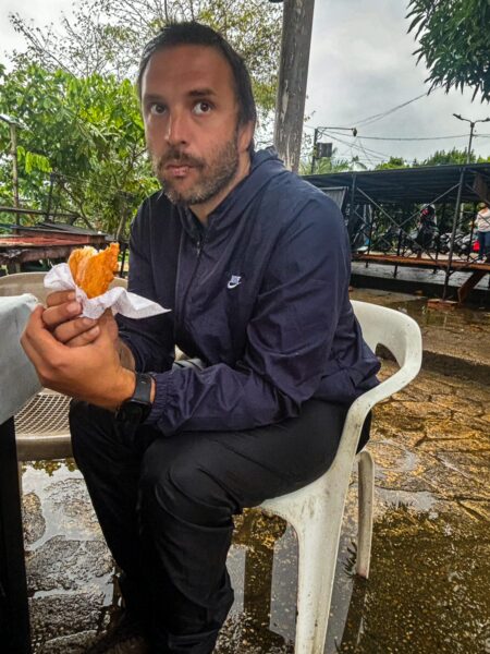 A man sitting on a white chair in Leticia, Amazonas, Colombia, holding a snack, surrounded by greenery and a cloudy sky.