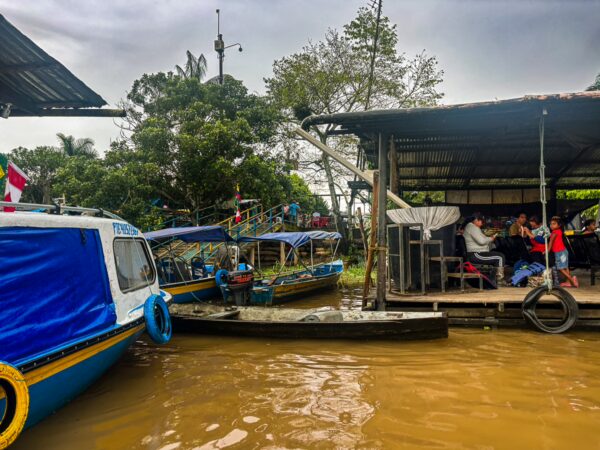Colorful boats docked at a riverbank in Leticia, Amazonas, Colombia, with people gathered under a shaded structure nearby.