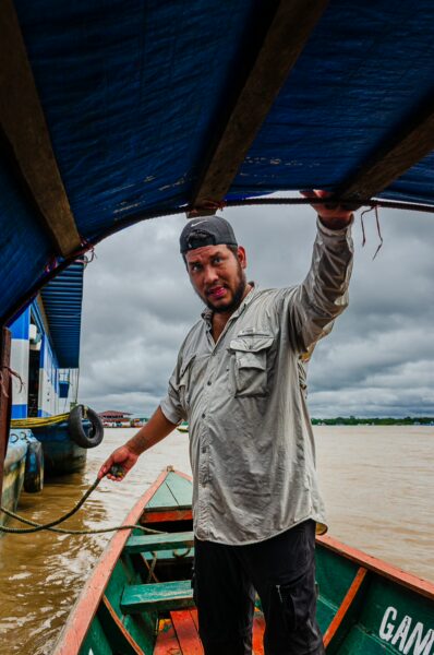 A man maneuvering a boat under a blue canopy on the Amazon River, surrounded by cloudy skies in Leticia, Amazonas, Colombia.