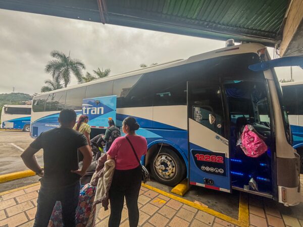 Travelers boarding a blue and white bus at a transport hub in Bucaramanga, with palm trees visible in the background.