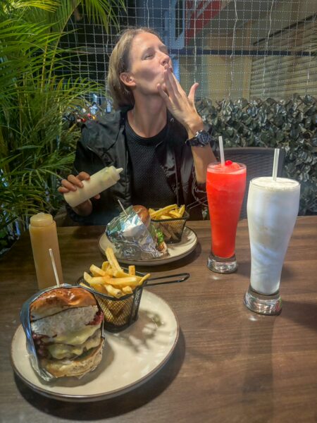 A diner enjoying a burger and fries in Bucaramanga, Colombia, with colorful drinks on the table and greenery in the background.