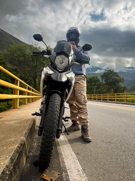 Motorbike rider standing beside a dual-sport bike on a mountain road bridge in Chicamocha Canyon, Colombia