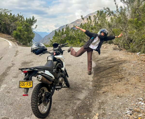 A person in a helmet playfully poses beside a motorcycle on a scenic road, surrounded by lush greenery in Chicamocha Canyon, Colombia.