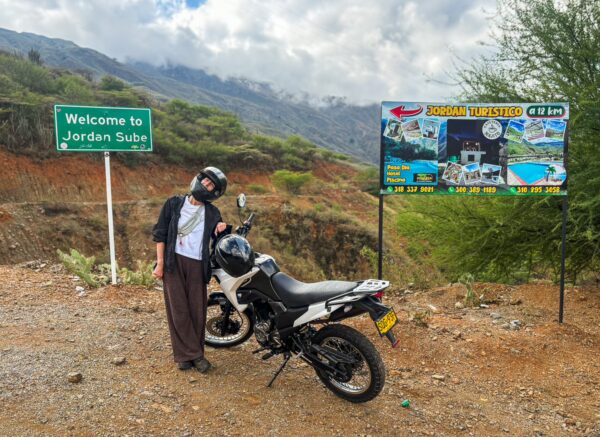 Motorbike rider standing beside a dual-sport bike at a roadside welcome sign for Jordan Sube in Chicamocha Canyon