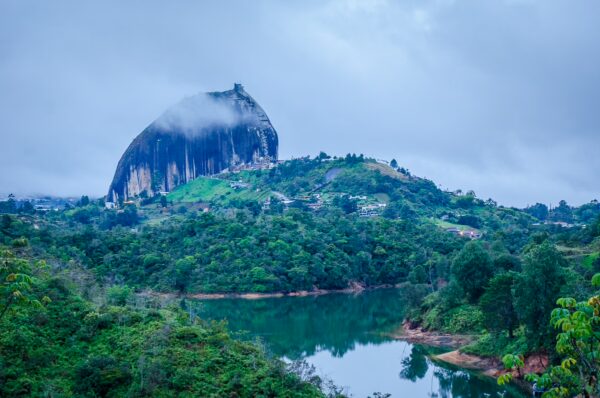 El Peñol Rock emerges majestically from lush green hills, reflecting in the calm waters of the lake below, under a cloudy sky.