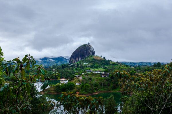 El Peñol Rock in Guatapé, Colombia, rises dramatically above lush green hills and a tranquil lake under a cloudy sky.