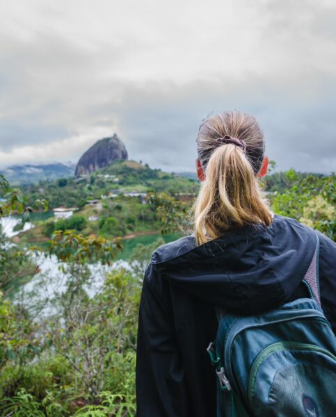 A person with a ponytail and backpack gazes at El Peñol Rock in Guatapé, Antioquia, surrounded by lush greenery and cloudy skies.