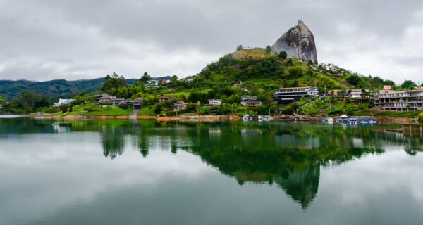 El Peñol Rock in Guatape, Antioquia, Colombia, rises over a tranquil lake, with lush greenery and quaint houses reflecting in the water.