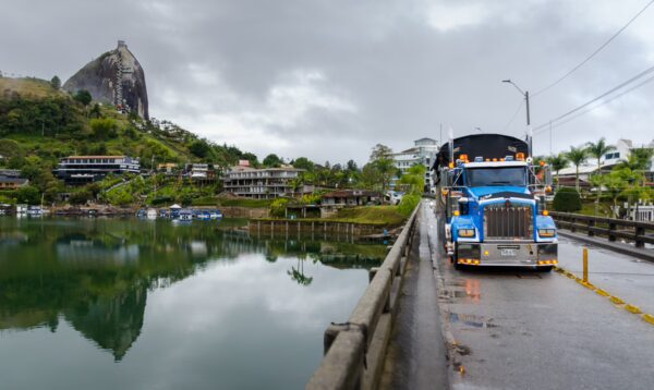 El Peñol Rock in Guatapé, Colombia, towers over a calm lake, with a blue truck parked on a bridge beside quaint houses and trees.