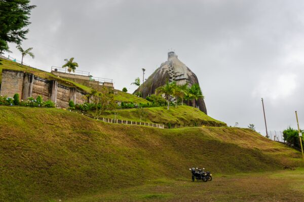 El Peñol Rock in Guatapé, Antioquia, Colombia, stands tall against a cloudy sky, surrounded by lush green hills and a grassy landscape.