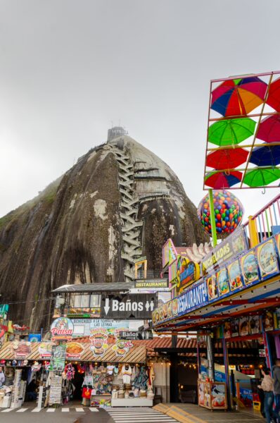 El Peñol Rock in Guatapé, Antioquia, Colombia, towers over colorful carnival stalls and a vibrant amusement area.