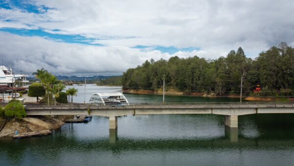 Guatapé, Antioquia: A serene view of the calm waters reflecting clouds, with a bridge connecting lush green banks and nearby boats.