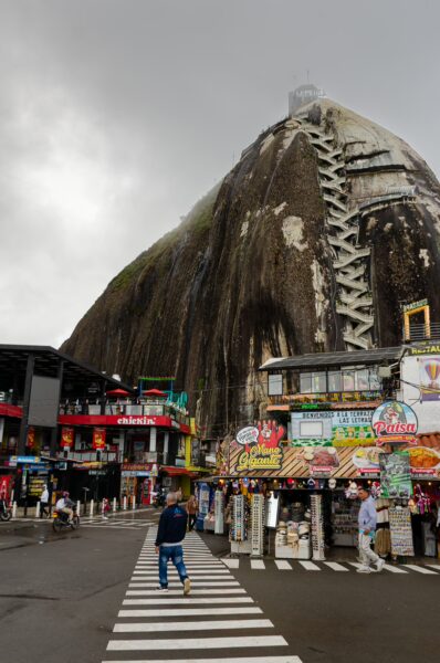 El Peñol Rock in Guatapé, Antioquia, Colombia, towers over colorful storefronts and a crosswalk under an overcast sky.
