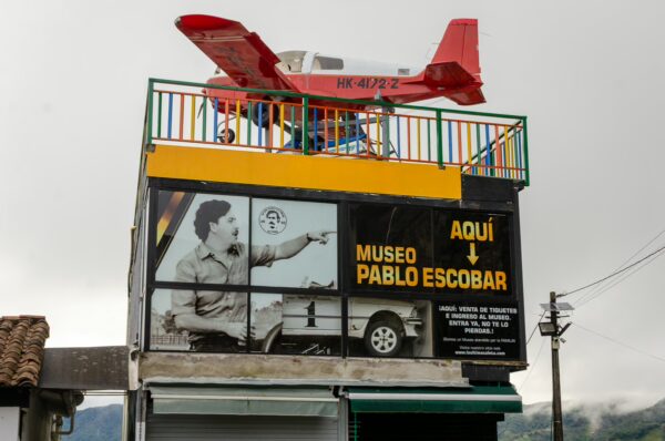 A red airplane model rests atop the Pablo Escobar Museum in Guatapé, Colombia, with vibrant signage and cloudy skies.