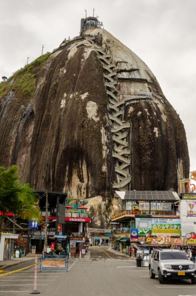El Peñol Rock in Guatapé, Colombia, towering over colorful shops and vehicles, under a cloudy sky with steep steps leading to the summit.