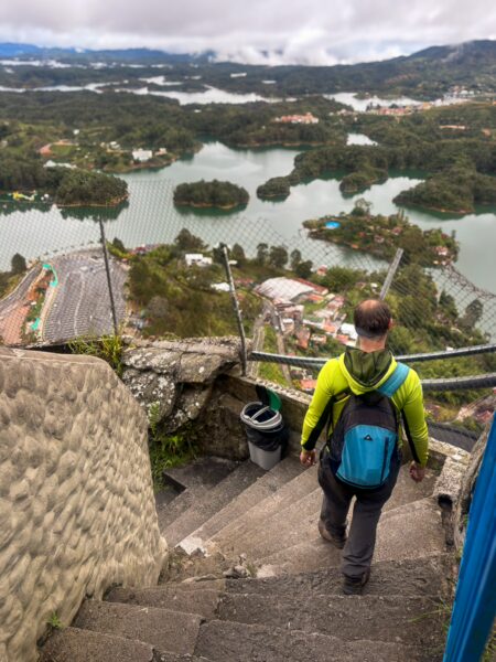El Peñol Rock, Guatapé, Antioquia, Colombia. A hiker descends stone steps, overlooking a lush lake and islands under a cloudy sky.