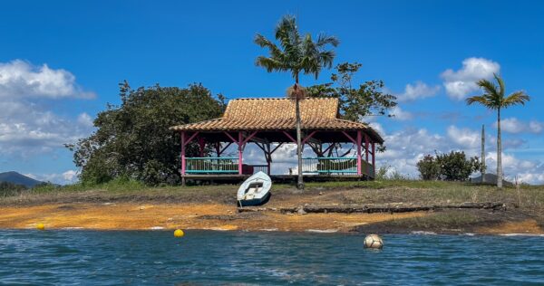 Guatapé Reservoir, Antioquia, Colombia features a vibrant waterfront hut under clear blue skies, surrounded by lush greenery.
