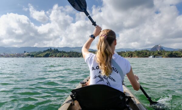 Kayaker paddling on the serene Guatapé Reservoir in Antioquia, Colombia, with vibrant green hills and clouds in the background.
