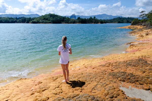 Guatapé Reservoir, Antioquia, Colombia: A woman stands on a vibrant orange shoreline with clear blue waters and lush green hills in the background.