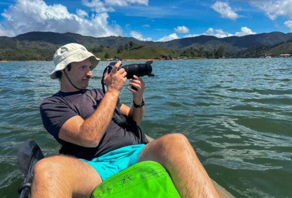 Kayaker capturing photos on the Guatapé Reservoir in Antioquia, Colombia, surrounded by lush green hills and a bright blue sky.
