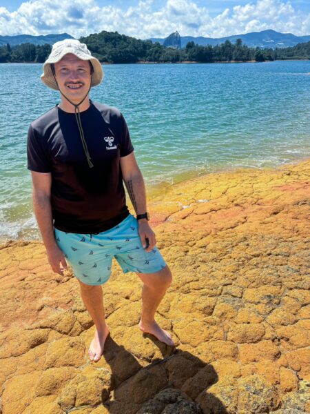 Guatapé Reservoir, Antioquia, Colombia: A person stands on vibrant orange and yellow rocks by the water, with lush greenery and mountains in the background.