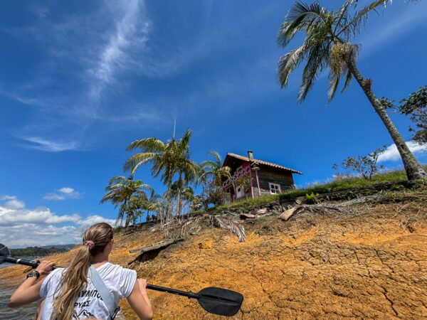 A woman paddles near the colorful shoreline of Guatapé Reservoir, Antioquia, Colombia, with lush palm trees and blue skies above.