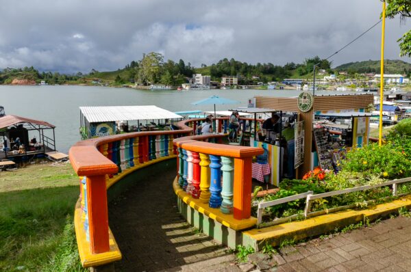 Colorful railing lines the path to the water in Guatapé, with vibrant shops and boats visible along the serene lake shore.